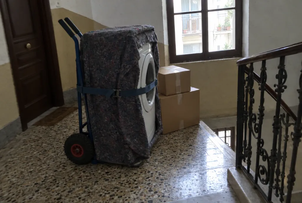 Lavadora blanca envuelta en una manta de mudanza, de pie sobre una carretilla de dos ruedas y sujeta con una cincha, en el rellano de un edificio antiguo con suelo de terrazo y barandilla de forja
