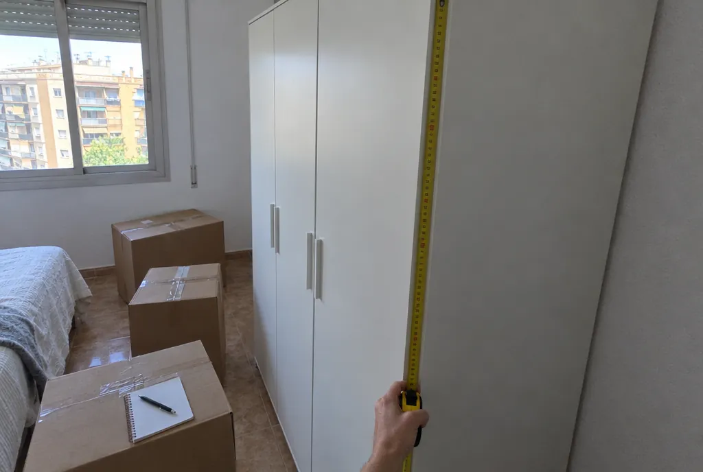 Mano sujetando un metro amarillo desplegado contra el lateral de un armario blanco alto en un dormitorio con cajas de cartón precintadas y una libreta encima de una de ellas