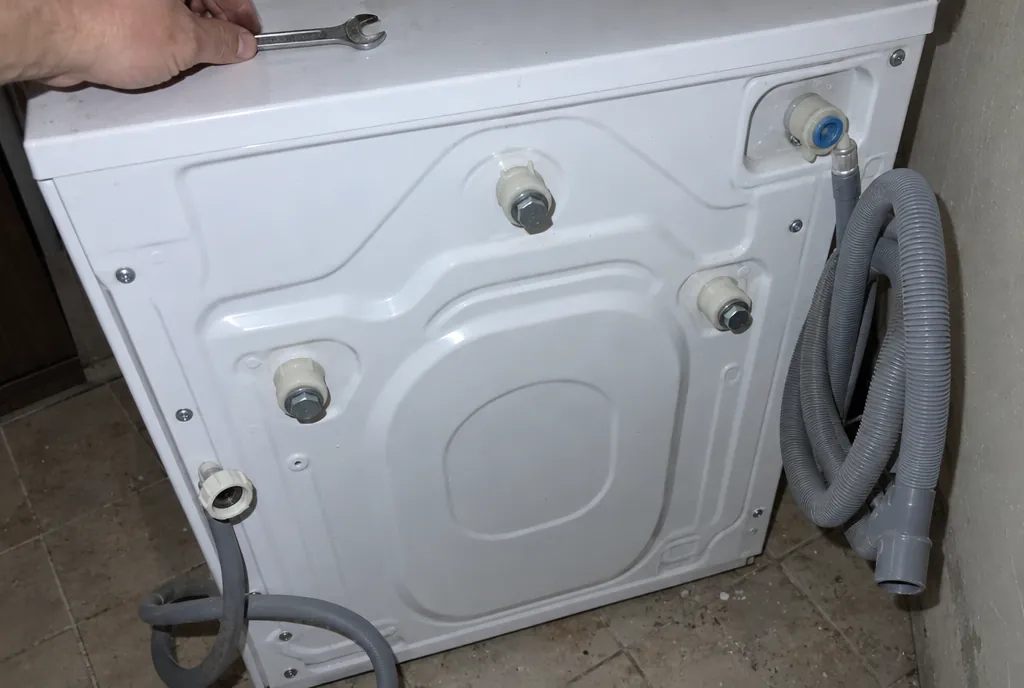 Trasera de una lavadora blanca separada de la pared con tres tornillos de transporte de cabeza hexagonal y sus casquillos de plástico colocados, una mano con una llave fija y las mangueras desconectadas